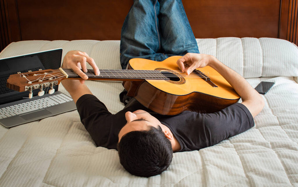 Mladý muž leží v posteli, hraje na kytaru a používá počítač, aby se učil doma. Volný čas doma: technologie a hudba. - Fotografie, Obrázek