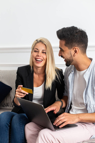 Приємна блондинка посміхається і читає кредитні картки посвідчення веселим етнічним хлопцям з ноутбуком, сидячи на дивані і роблячи онлайн покупки разом - Фото, зображення
