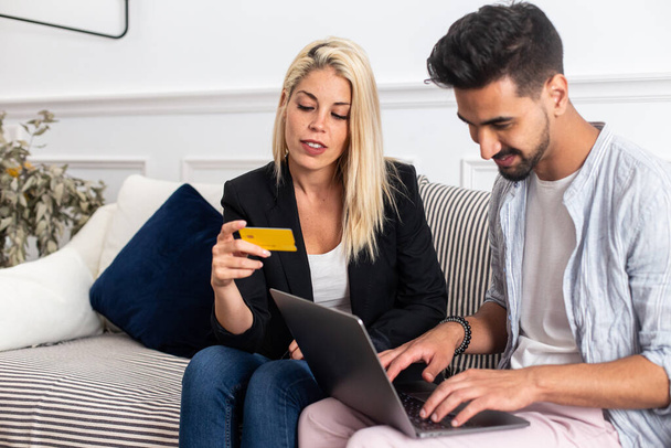 Восхитительная блондинка, улыбающаяся и читающая удостоверения кредитной карты веселому этническому парню с ноутбуком, сидящему на диване и совершающему совместные покупки в интернете - Фото, изображение