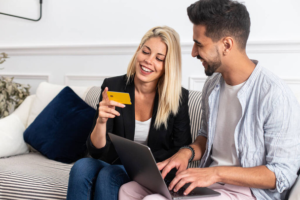 Приємна блондинка посміхається і читає кредитні картки посвідчення веселим етнічним хлопцям з ноутбуком, сидячи на дивані і роблячи онлайн покупки разом. - Фото, зображення