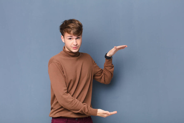 young blonde man holding an object with both hands on side copy space, showing, offering or advertising an object isolated against flat wall - Foto, afbeelding