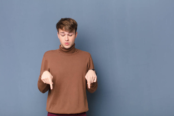 young blonde man with open mouth pointing downwards with both hands, looking shocked, amazed and surprised isolated against flat wall - Foto, Bild