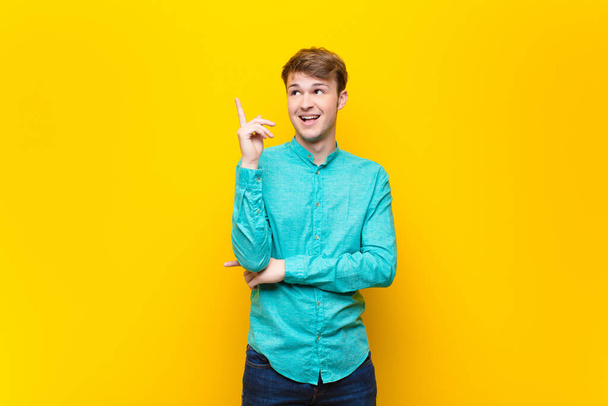young blonde man smiling happily and looking sideways, wondering, thinking or having an idea isolated against flat wall - Photo, image