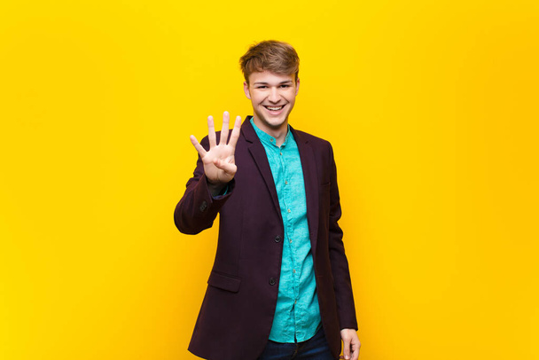 young blonde man smiling and looking friendly, showing number four or fourth with hand forward, counting down isolated against flat wall - Foto, Imagem