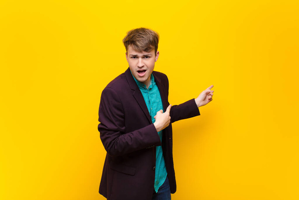 young blonde man feeling shocked and surprised, pointing to copy space on the side with amazed, open-mouthed look isolated against flat wall - Фото, зображення