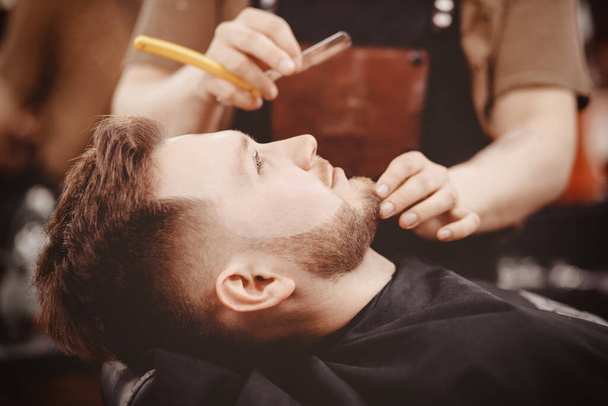 Мужчина парикмахер бреет бороду клиента на стуле Парикмахерской
 - Фото, изображение