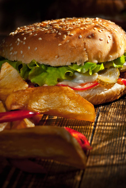 Burger with french fries on a dark wooden board. Cheeseburger with cutlet and herbs on black background. Food with contrasting dramatic light. Potatoes with red peppers closeup. - Photo, image
