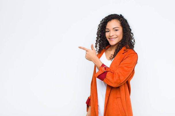 young black woman looking excited and surprised pointing to the side and upwards to copy space against white wall - Foto, Imagem