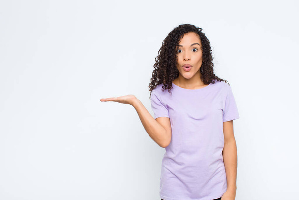 young black woman looking surprised and shocked, with jaw dropped holding an object with an open hand on the side against white wall - Foto, Imagem