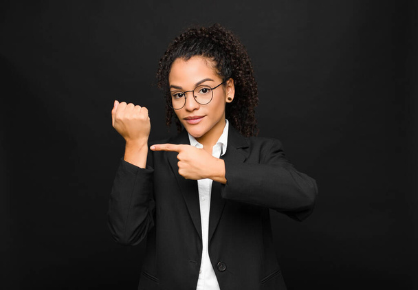 young black woman looking impatient and angry, pointing at watch, asking for punctuality, wants to be on time against black wall - Фото, изображение