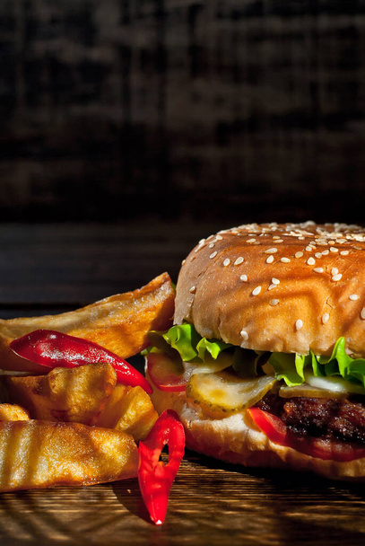Burger with french fries on a dark wooden board. Cheeseburger with cutlet and herbs on black background. Food with contrasting dramatic light. Potatoes with red peppers closeup. - Foto, Bild