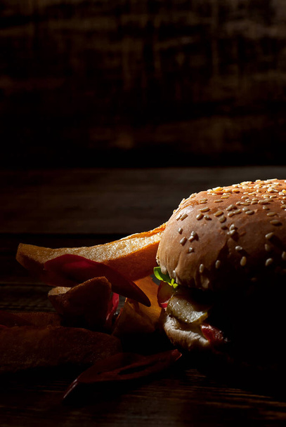 Burger with french fries on a dark wooden board. Cheeseburger with cutlet and herbs on black background. Food with contrasting dramatic light. Potatoes with red peppers closeup. - Φωτογραφία, εικόνα