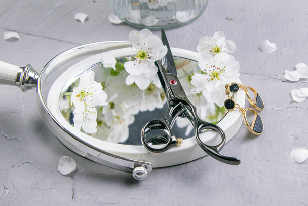 Cosmetic set on grey dressing table. Top views, close-up. Scissors and earrings stand on a mirror sprinkled with white flowers. - Photo, Image