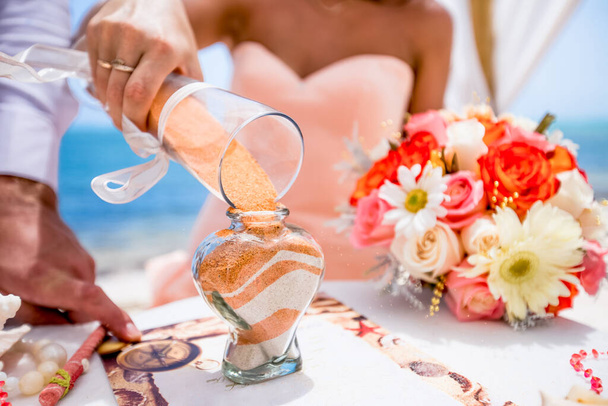 Bride and groom pouring colorful different colored sands into the crystal vase close up during symbolic nautical decor destination wedding marriage ceremony on sandy beach in front of the ocean in Punta Cana, Dominican republic - Foto, afbeelding