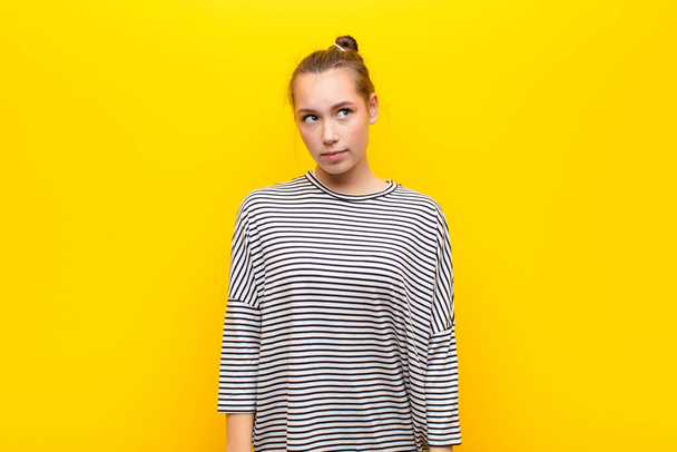 young blonde pretty girl with a worried, confused, clueless expression, looking up to copy space, doubting against yellow wall - Foto, Bild