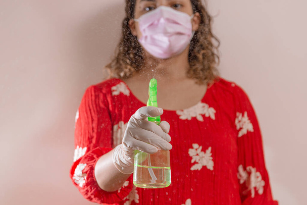 Mujer latina con vestido rojo tomando precauciones por el Covid-19 usando una mascarilla, guantes y un atomizador con alcohol para prevenir infeccion del coronavirus en el 2020 - Photo, Image