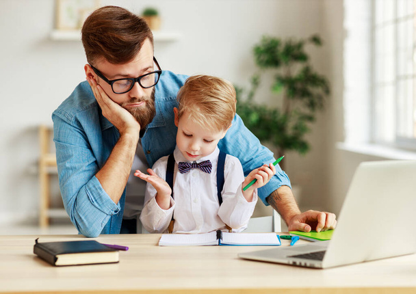 Kafası karışmış küçük bir çocuk ve babası masada oturuyor ve zor bir görev üzerinde düşünürken çevrimiçi çalışıyor ve evde ödevlerini birlikte yapıyorlar. - Fotoğraf, Görsel