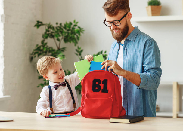 Boldog kisfiú apával másoló könyveket tesz a hátizsákba, miközben otthon áll az asztal mellett, és készül az iskolára. - Fotó, kép