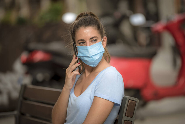 Девушка пользуется телефоном на улице в медицинской маске во время коронавирусного карантина
 - Фото, изображение