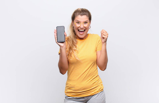 young pretty latin woman feeling shocked, excited and happy, laughing and celebrating success, saying wow! with a smartphone against white wall - Foto, Bild