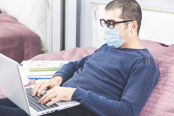 Korunaklı maskeli bir adam Coronavirus Covid-19 salgınında karantina sırasında evden çalışıyor. Dizüstü bilgisayarla yatakta oturuyor. Ev kavramından çalış. - Fotoğraf, Görsel