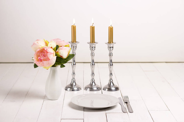 Is klaar voor het feest. Witte houten tafel geserveerd met bord en toestel met kaarsen en een vaas bloemen. Focus op de plaat. - Foto, afbeelding