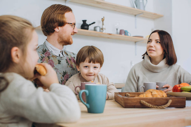 Jeune famille heureuse avec deux enfants prenant le petit déjeuner ensemble. rousse papa, les enfants et maman brune ensemble dans la cuisine
. - Photo, image
