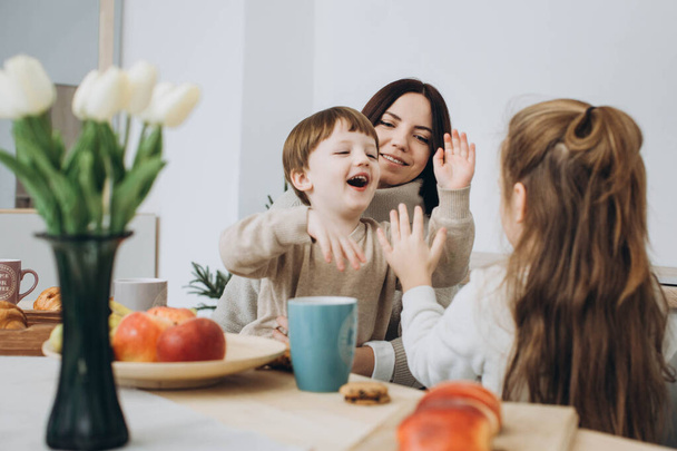 Νεαρή ευτυχισμένη οικογένεια με δύο παιδιά που τρώνε πρωινό μαζί. κοκκινομάλλης μπαμπάς, παιδιά και μελαχρινή μαμά μαζί στην κουζίνα. - Φωτογραφία, εικόνα