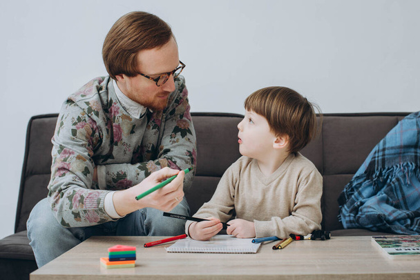 Gözlüklü kızıl saçlı baba ve küçük oğlu renkli kalemler çiziyorlar, babalarını seviyorlar ve küçük sevimli bir okul çocuğu birlikte eğleniyorlar.. - Fotoğraf, Görsel