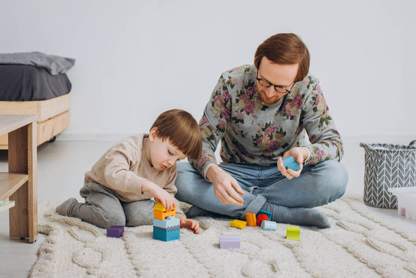 Father in glasses and adorable toddler son building wooden constructor tower. - Foto, afbeelding