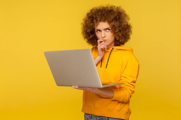 Confused puzzled thoughtful woman with fluffy curly hair in urban style hoody holding laptop thinking over difficult question, pondering solution. indoor studio shot isolated on yellow background - Foto, Imagem