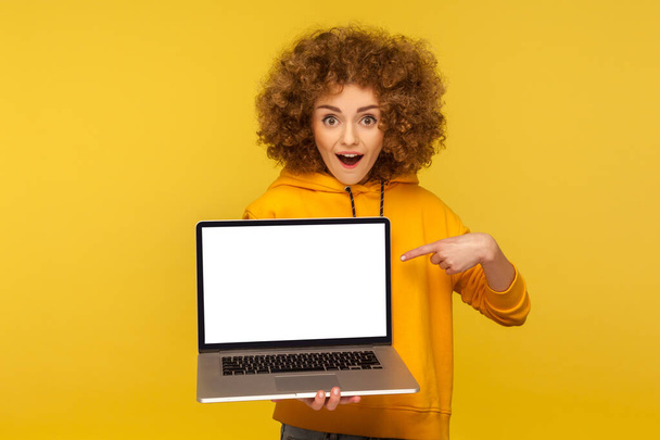 Shocked, surprised woman with fluffy curly hair in urban style hoody pointing at empty laptop screen and looking with amazement, showing mock up, blank display for web ad. indoor studio shot isolated - Φωτογραφία, εικόνα