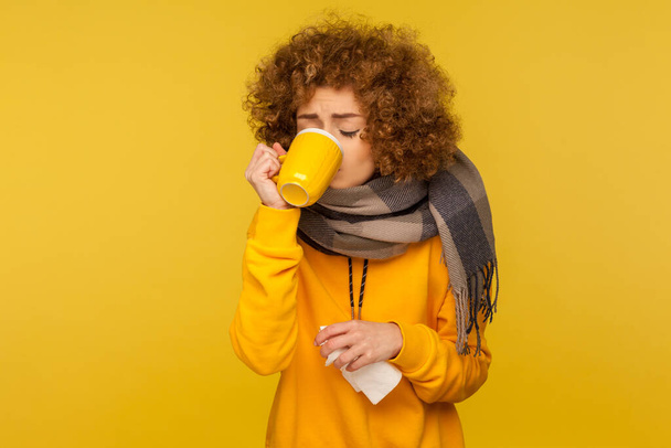 Influenza treatment. Flu-sick woman with curly hair, wrapped in warm scarf, drinking hot tea and holding napkin, taking pills, treating fever with medicine. indoor studio shot, yellow background - Фото, зображення