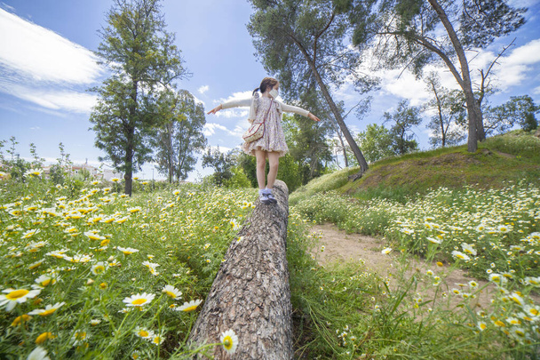 Uma menina a andar num tronco no meio do campo da margarida. Ela usa uma máscara facial.
 - Foto, Imagem