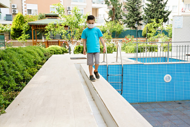 Chlapec s lékařskou maskou chodí kolem prázdného bazénu. Koncept léta2020. Žádný cestovní ruch  - Fotografie, Obrázek