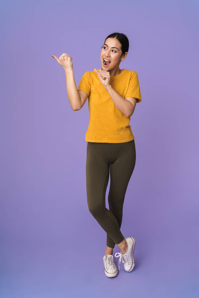 Full length of a cheerful young pretty sportswoman standing isolated over violet background, presenting copy space - Photo, image