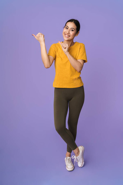 Full length of a cheerful young pretty sportswoman standing isolated over violet background, presenting copy space - Фото, изображение