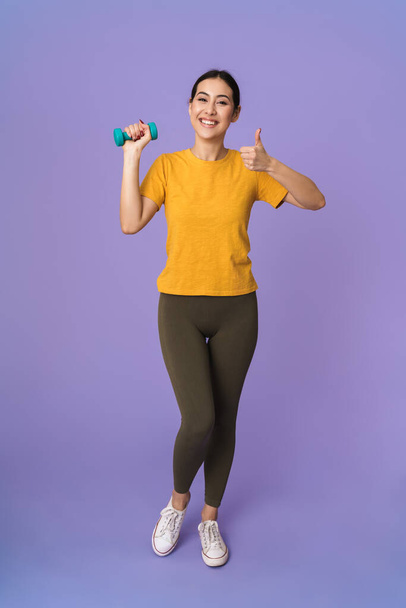 Full length of a cheerful young pretty sportswoman standing isolated over violet background, lifting dumbbell, showing thumbs up - Foto, Imagen