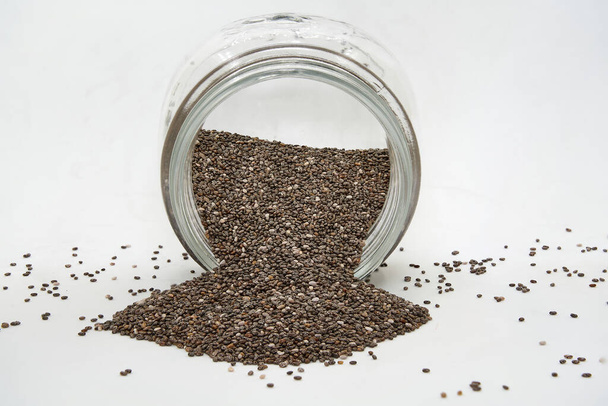 Lidless glass pot lying full of chia seeds that have spilled over a white background - Photo, Image