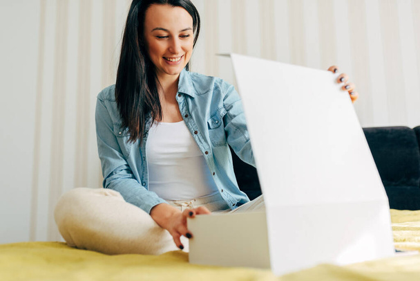 Horizontal indoor image of smiling young woman sitting on the bed and unpacking the parcel. Surprised female receive a gift box for her birthday at home. Online internet commerce delivery concept - Fotografie, Obrázek