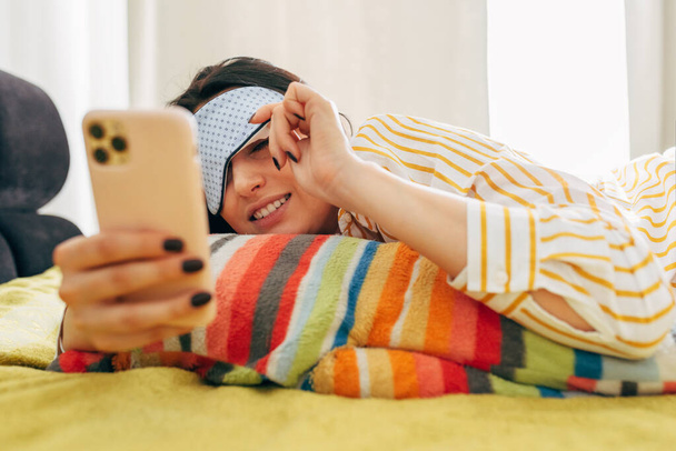 Awake young woman lying on the bed with sleep mask, looking to mobile phone screen, reading messages on it. Female freelancer wearing striped shirt sends message on smartphone before resting at home.  - 写真・画像