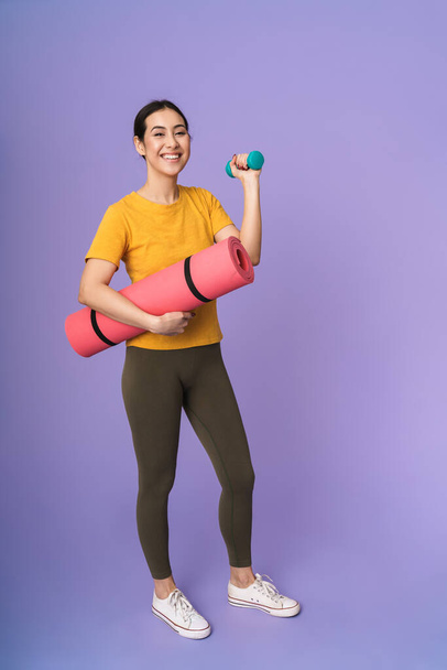 Full length of a cheerful young pretty sportswoman standing isolated over violet background, carrying fitness mat while lifting dumbbell - Foto, Imagen