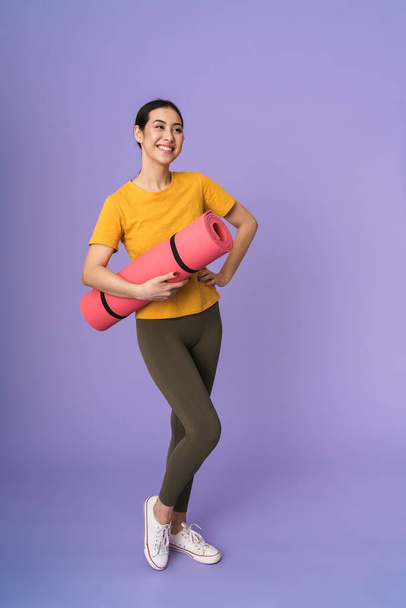 Full length of a cheerful young pretty sportswoman standing isolated over violet background, carrying fitness mat - Foto, Imagen