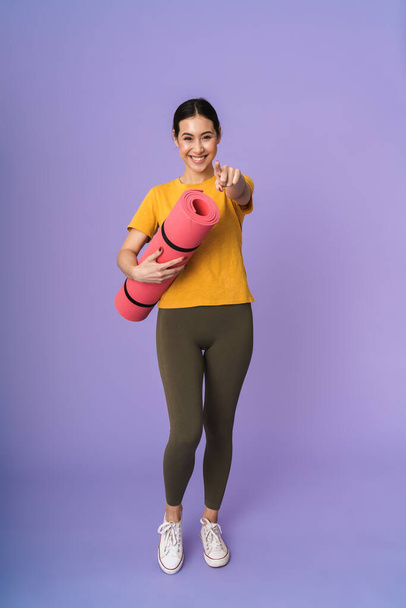 Full length of a cheerful young pretty sportswoman standing isolated over violet background, carrying fitness mat, pointing finger at camera - Фото, зображення