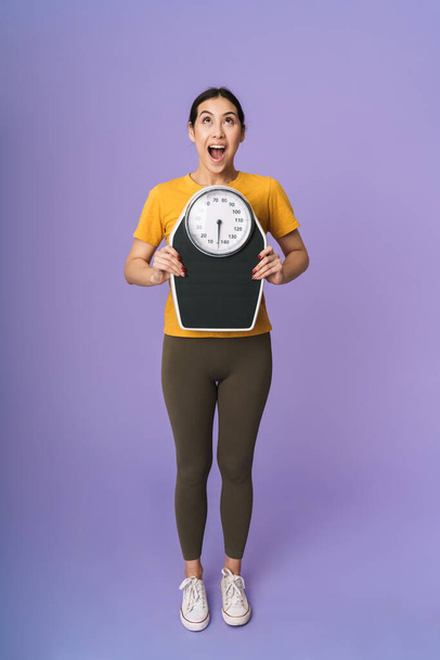 Full length of a cheerful excited young pretty sportswoman standing isolated over violet background, showing scales - Fotoğraf, Görsel