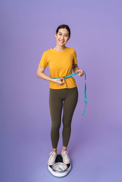 Full length of a cheerful young pretty sportswoman standing on scales isolated over violet background, holding measuring tape around her waist - Fotó, kép
