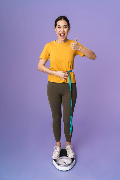 Full length of a cheerful young pretty sportswoman standing on scales isolated over violet background, holding measuring tape around her waist, thumbs up - Foto, Imagen