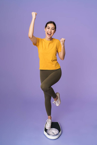 Full length of a cheerful young pretty sportswoman standing on scales isolated over violet background, celebrating - 写真・画像