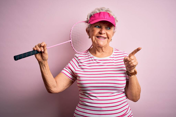 Старшая красивая спортсменка держит бадминтон ракетку на изолированном розовом фоне очень счастливо указывая рукой и пальцем в сторону
 - Фото, изображение