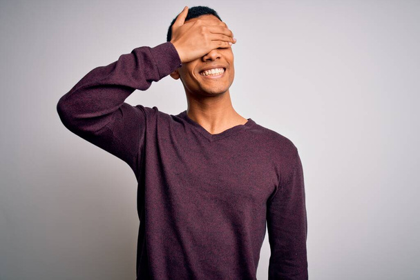 Jeune bel homme afro-américain portant un pull décontracté sur fond blanc souriant et riant avec la main sur le visage couvrant les yeux pour la surprise. Concept aveugle
. - Photo, image